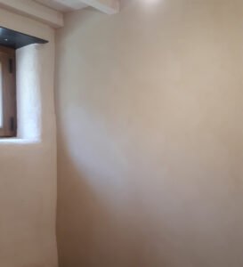 Mur intérieur enduit dans une teinte sable douce autour d’une petite fenêtre en bois, finition mate nuagée réalisée par Décors & Matières.
